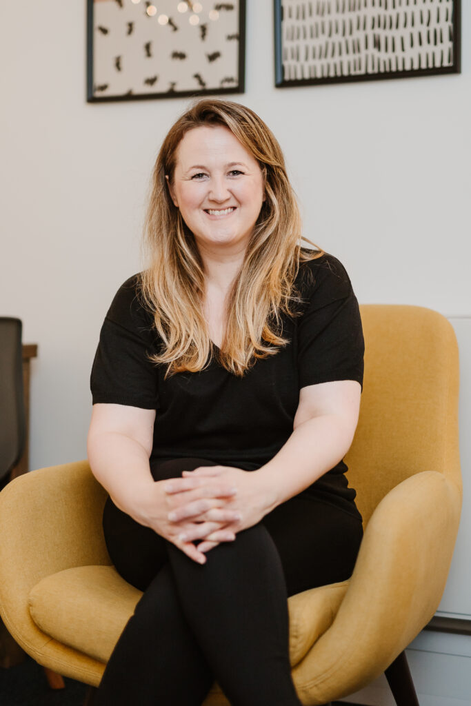 Woman smiles while sitting in therapy space on mustard coloured cosy chair.  Image is tightly cropped in portrait orientation.
