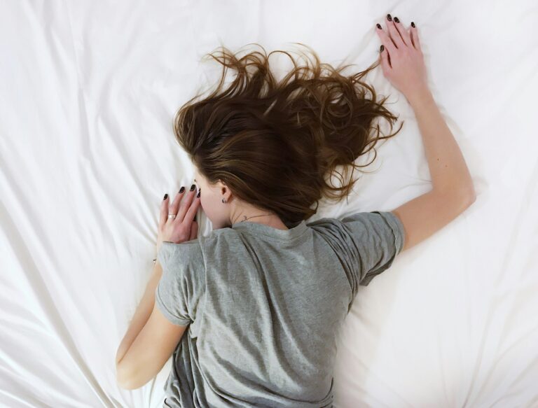 Woman in grey t-shirt lies face down with left arm spread out and face buries into white sheet